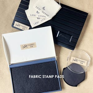 May include: A black rectangular stamp pad with a white lid, a smaller circular black stamp pad with a clear lid, and a black grooved base. The items are labelled Magic Fabric Ink. Three off-white fabric tags with the text Lexi Lynne are included.