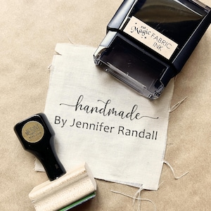 May include: A black self-inking stamp labeled PM Magic Fabric Ink and a wooden handle stamp with a black top, both resting on a piece of off-white fabric printed with the text handmade By Jennifer Randall in a black script and sans-serif font.