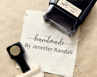 May include: A black self-inking stamp labeled PM Magic Fabric Ink and a wooden handle stamp with a black top, both resting on a piece of off-white fabric printed with the text handmade By Jennifer Randall in a black script and sans-serif font.