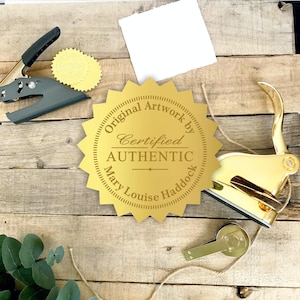 May include: A gold-colored starburst-shaped seal with the text Original Artwork by Certified AUTHENTIC MARY LOUISE HADDOCK, accompanied by a gold metal embosser, a dark gray handheld hole punch, and a small gold metal component.
