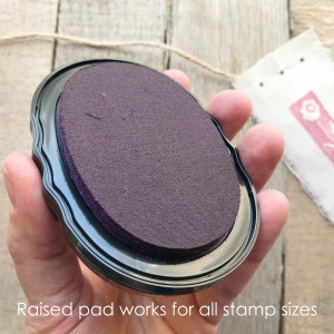 Peut inclure: Tampon encreur noir avec une surface d'encre violette ovale et sur&eacute;lev&eacute;e. Le texte "Raised pad works for all stamp sizes" est visible en bas. Le tampon encreur est tenu dans une main, avec une petite &eacute;tiquette en tissu estamp&eacute;e en arri&egrave;re-plan.
