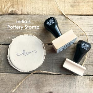 May include: A pottery stamp with the initials "hw" imprinted on a round, light-colored clay piece. Two additional stamps with black handles and wooden blocks are visible. The text "Initials Pottery Stamp" is in the upper left corner. A natural twine is also present.