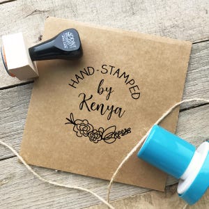 May include: A brown paper envelope with the text "HAND-STAMPED by Kenya" in black script, along with a floral design. Two rubber stamps, one black and one blue, are positioned near the envelope. A length of twine is also present.
