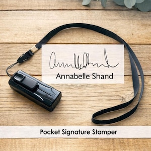 May include: A black pocket signature stamper with a lanyard. The stamper has the name "Annabelle Shand" printed on it. The words "Pocket Signature Stamper" are also visible. The background is a wooden surface.