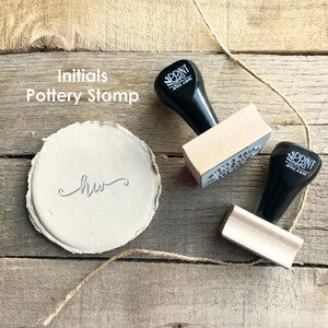 Puede incluir: Un juego de sellos de cerámica con el texto "Initials Pottery Stamp" y una muestra de las iniciales "hw". El juego incluye dos sellos con mangos negros y bloques de madera, y una pieza redonda de arcilla con las iniciales estampadas. El fondo es una superficie de madera.