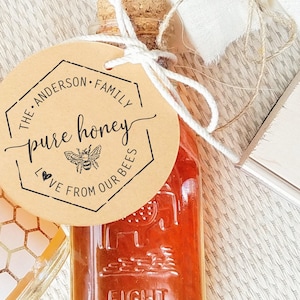 A hexagon graphic with a bee and text inside it  is stamped in ink on a paper label tied to a glass bottle full of honey. The center text reads Raw Honey. At the top is a family name and below reads Love from our bees.