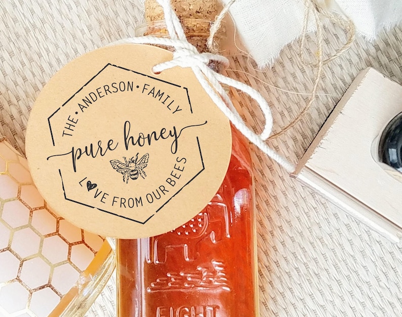 A hexagon graphic with a bee and text inside it  is stamped in ink on a paper label tied to a glass bottle full of honey. The center text reads Raw Honey. At the top is a family name and below reads Love from our bees.