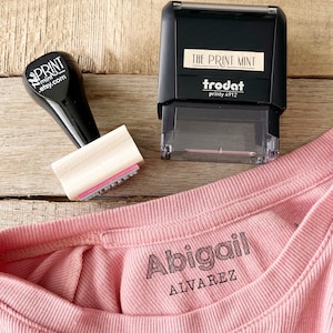 May include: A black stamp with the text "THE PRINT MINT" and a smaller stamp with the text "PRINT mint .etsy.com" are next to a pink shirt with the name "Abigail" and the last name "ALVAREZ" printed on it.