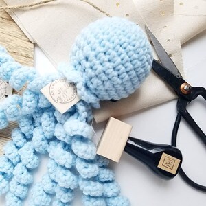 Peut inclure: Un jouet poulpe crochet&eacute; bleu clair avec une t&ecirc;te ronde et des tentacules boucl&eacute;es. Le poulpe porte une &eacute;tiquette avec l'inscription "An Original Creation". Un tampon &agrave; manche noir et des ciseaux sont &eacute;galement dans l'image.