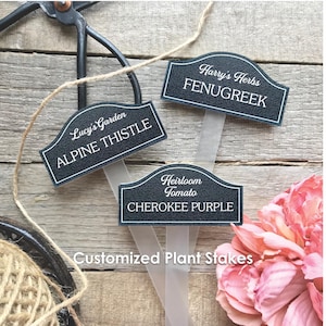 May include: Three black and white plant labels with text for Alpine Thistle, Fenugreek, and Cherokee Purple. The labels are on a wooden surface with gardening tools and twine. The text "Customized Plant Stakes" is at the bottom.