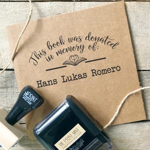 May include: A kraft paper book donation card with the text "This book was donated in memory of: Hans Lukas Romero." A black stamp and a stamp pad are next to the card. The stamp says "THE PRINT MINT" and "trodat printy 4912".