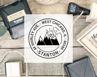 May include: A black and white custom address stamp with the text "44 SHIRLEY AVE. - WEST CHICAGO, IL - 60185 - STANTON" encircling a mountain range design with a heart. The stamp is surrounded by other stamps and twine.