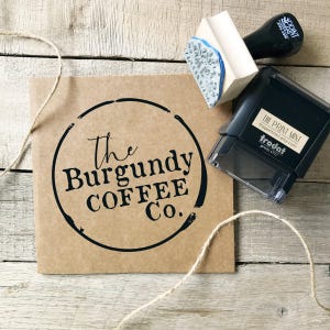 May include: A kraft paper bag with the text "The Burgundy Coffee Co." in a black circle. A wooden stamp with a floral design and a black handle, and a black self-inking stamp are next to the bag. Twine is on the wooden surface.