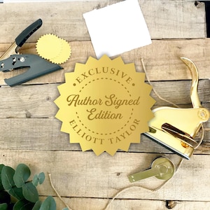 May include: A gold foil seal with a serrated edge featuring the text "EXCLUSIVE Author Signed Edition ELLIOTT TAYLOR" in the center. The seal is positioned between a dark gray metal hand embosser and a polished gold metal hand embosser.