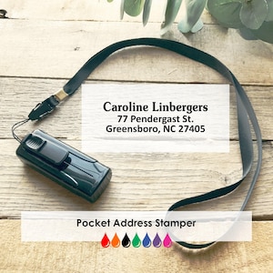 May include: A black pocket address stamper with a black lanyard. The stamper has the text "Caroline Linbergers, 77 Pendergast St. Greensboro, NC 27405" and "Pocket Address Stamper". The stamper has a row of colorful ink drop icons.