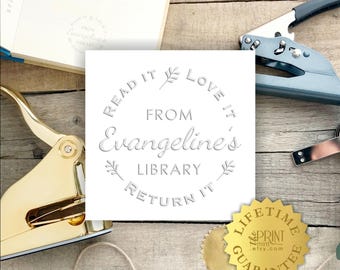 May include: A square white paper insert featuring an embossed circular design with the text "READ IT LOVE IT FROM Evangeline's LIBRARY RETURN IT" and small leaf icons. The insert is surrounded by a gold metal embosser, a blue tool, and a gold seal.