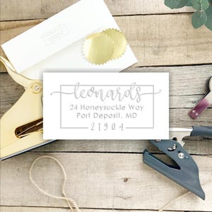 May include: A rectangular white address label featuring the name Leonards in a cursive script, followed by the address 24 Honeysuckle Way, Port Deposit, MD 21904, all enclosed within a thin rectangular border, positioned near gold embossing tools.