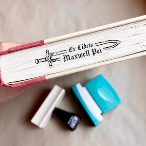 Puede incluir: Un libro con bordes rojos y un sello personalizado que dice "Ex Libris Maxwell Pei" con una ilustración de espada. Se ven varias almohadillas de sellos, incluyendo blanco, negro y turquesa.