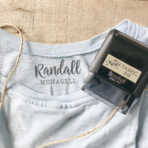May include: A pale blue t-shirt with the name "Randall Monacell" printed on the front. A black "Magic Fabric Ink" stamp is next to the shirt. A length of twine is draped across the shirt.