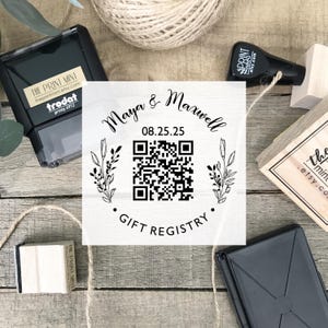 Può includere: Un timbro quadrato con il testo "Maya & Maxwell" e la data "08.25.25" con un codice QR e le parole "Gift Registry". Il timbro è circondato da elementi floreali. Sono visibili anche altri timbri e spago.
