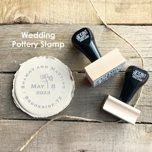 May include: A wedding pottery stamp with a black handle and wooden base, next to a stamped pottery piece. The stamp reads "SHAWNY AND MARTINO, MAY 8, 2023, BROOKSIDE, TX." The stamp is on a wooden surface.