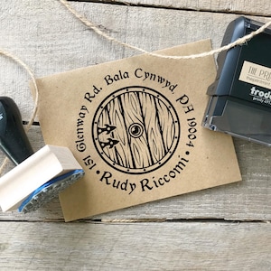 May include: A brown kraft paper envelope with a black circular stamp featuring a wooden shield design. The stamp includes the address "151 Glenway Rd. Bala Cynwyd, PA 19004" and the name "Rudy Riccomi". A black and a wooden stamp are also visible.