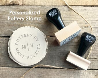 May include: Two black and light wood pottery stamps with the text "the print mint .etsy.com" on the handles, positioned next to a circular clay disc embossed with the text "POTTERY BY MATT VOLETTA" and the initials "M | V" in the center.
