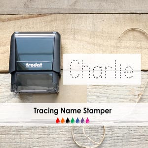 May include: A black Trodat tracing name stamper next to a paper with the name "Charlie" in dotted lines. The text "Tracing Name Stamper" is below the paper, with colorful ink drop icons. The background is a light brown wooden surface.
