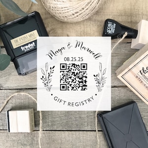 May include: A square stamp with the text "Maya & Maxwell" and a QR code, surrounded by floral elements. The stamp also includes the date 08.25.25 and the words "Gift Registry". Other stamps and twine are also visible.