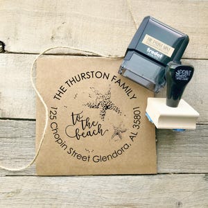 May include: A custom stamp with the text "The Thurston Family" and "to the beach" in a circular design, along with an address. A black ink pad and a stamp are next to the stamp. The stamp is on a brown paper.