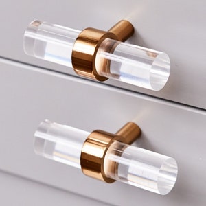 May include: Two clear acrylic cabinet pulls with gold-toned metal accents. The cylindrical pulls are mounted on a light gray cabinet. The hardware has a modern, minimalist design.