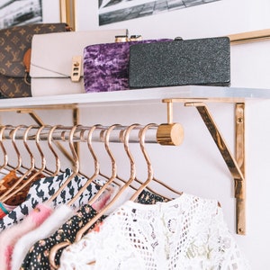 Lucite shelf with brass brackets, holding a clear acrylic rod with matching hardware, used to hang clothes. A variety of clutches and purses are displayed on the top of the shelf.