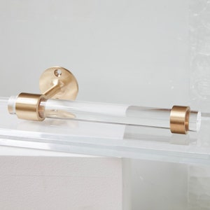 A clear lucite tubular rod fitted in two brass rings on both ends. One ring is attached to mounting hardware to install on a wall for the item to be used as a toilet paper holder. The hardware is solid brass, finished in satin brass.