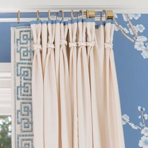 Beige drapes with a blue greek key trim are hung from a clear lucite curtain rod with polished brass hardware and matching curtain rings, installed in front of a blue floral wallpapered wall.