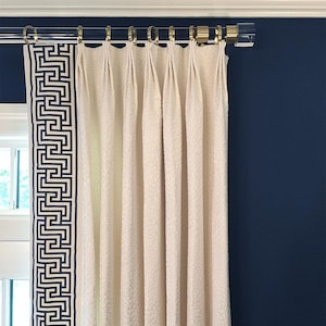 Clear round lucite curtain rod with polished brass hardware holding up beige drapery with Greek key style trim. Finials are lucite cylinder end caps.