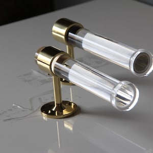 Two transparent glass tubes with brass bases are placed on a white surface, creating a minimalist and elegant design.