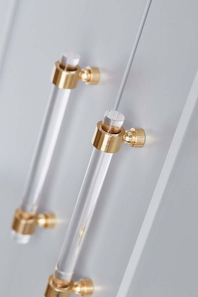 Set of clear lucite cabinet door handles or pulls with a crown post hardware base, finished in polished brass. The pair of pulls are installed on light grey cabinetry.