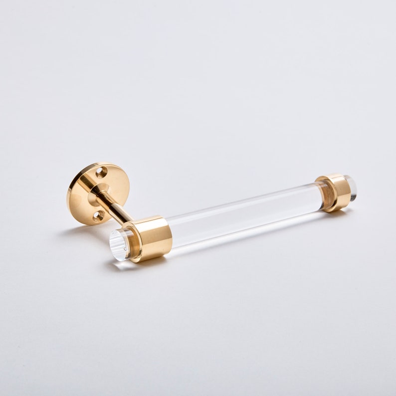 A clear lucite tubular rod fitted in two brass rings on both ends. One ring is attached to mounting hardware to install on a wall for the item to be used as a toilet paper holder. The hardware is solid brass, finished in polished brass.