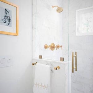 A lucite and satin brass towel bar is installed on the outside wall of a glass shower in a white bathroom. All of the shower accessories are brass to match.