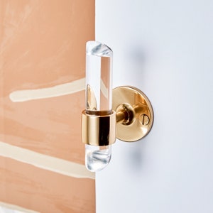 Cylindrical clear lucite piece with angled ends, inserted in a brass ring and wall mounted to be used as a robe or towel hook. Finish shown is gold polished brass.