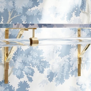 A clear lucite acrylic shelf with polished brass brackets attached to a blue foliage wallpapered wall. A single acrylic rod is attached with matching brass hardware for hanging clothes beneath the shelf.