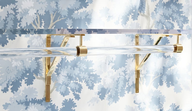 A clear lucite acrylic shelf with polished brass brackets attached to a blue foliage wallpapered wall. A single acrylic rod is attached with matching brass hardware for hanging clothes beneath the shelf.