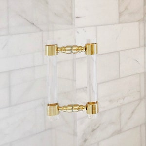 May include: A pair of clear acrylic door handles with gold metal accents. The handles are mounted on a white tiled wall.