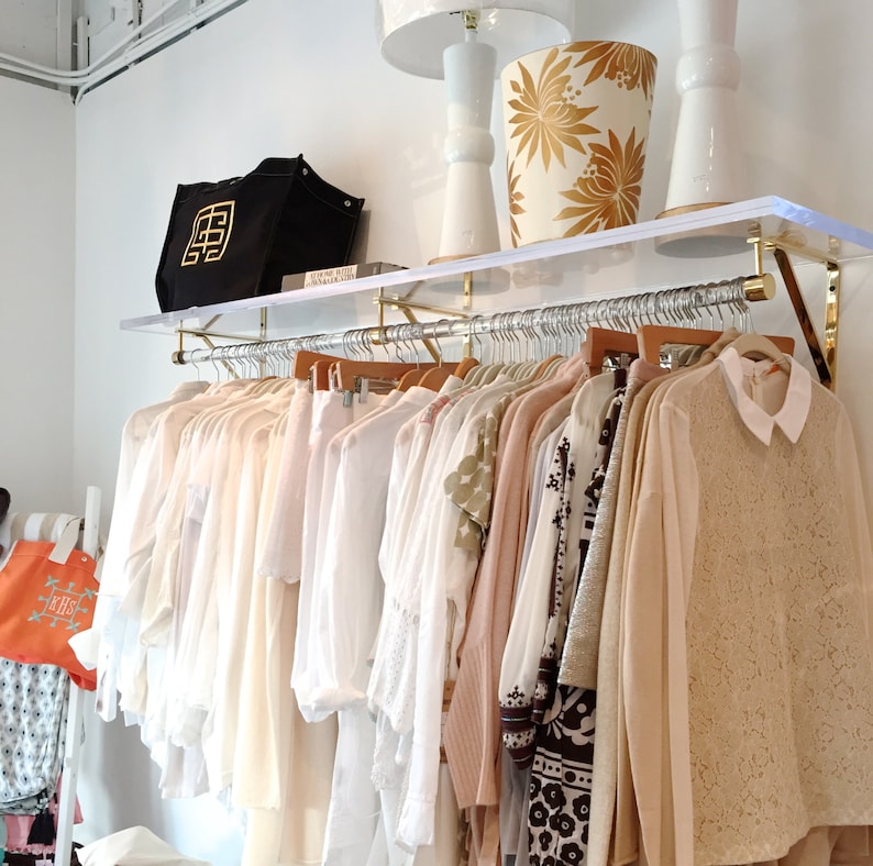 Lucite shelf with brass brackets, holding a clear acrylic rod with matching hardware, used to hang clothes in a retail space. Two lamps, a decorative trash can, and a bag are resting on top of the acrylic shelf.