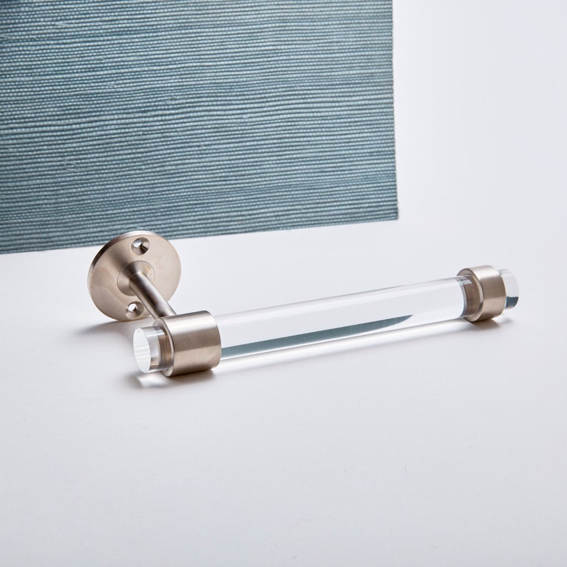 A clear lucite tubular rod fitted in two brass rings on both ends. One ring is attached to mounting hardware to install on a wall for the item to be used as a toilet paper holder. The hardware is solid brass, finished in satin nickel.