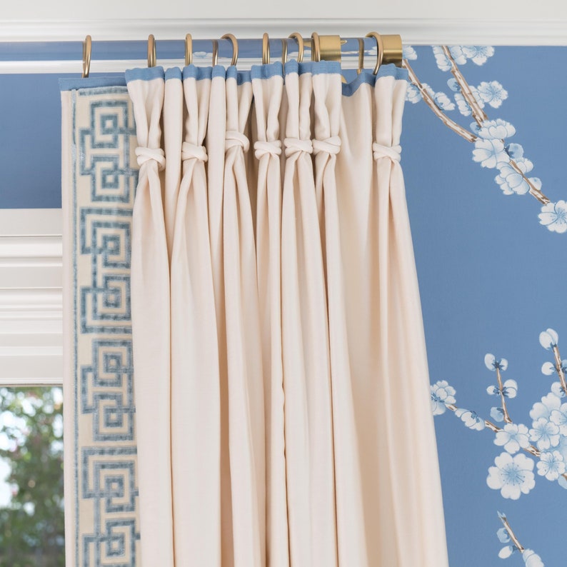 Beige drapes with a blue greek key trim are hung from a clear lucite curtain rod with polished brass hardware and matching curtain rings, installed in front of a blue floral wallpapered wall.