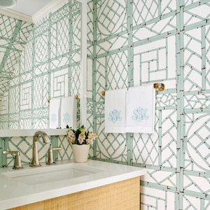 A bathroom with lattice style wallpaper features a simple vanity, large mirror, and lucite and brass towel rod, holding two white monogrammed towels.