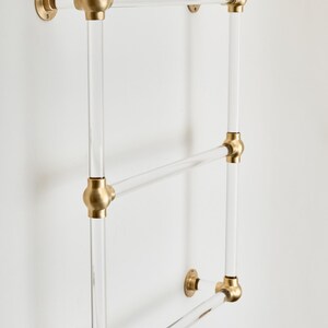 A clear glass towel rack with three horizontal bars and brass-colored hardware attached to a white wall.
