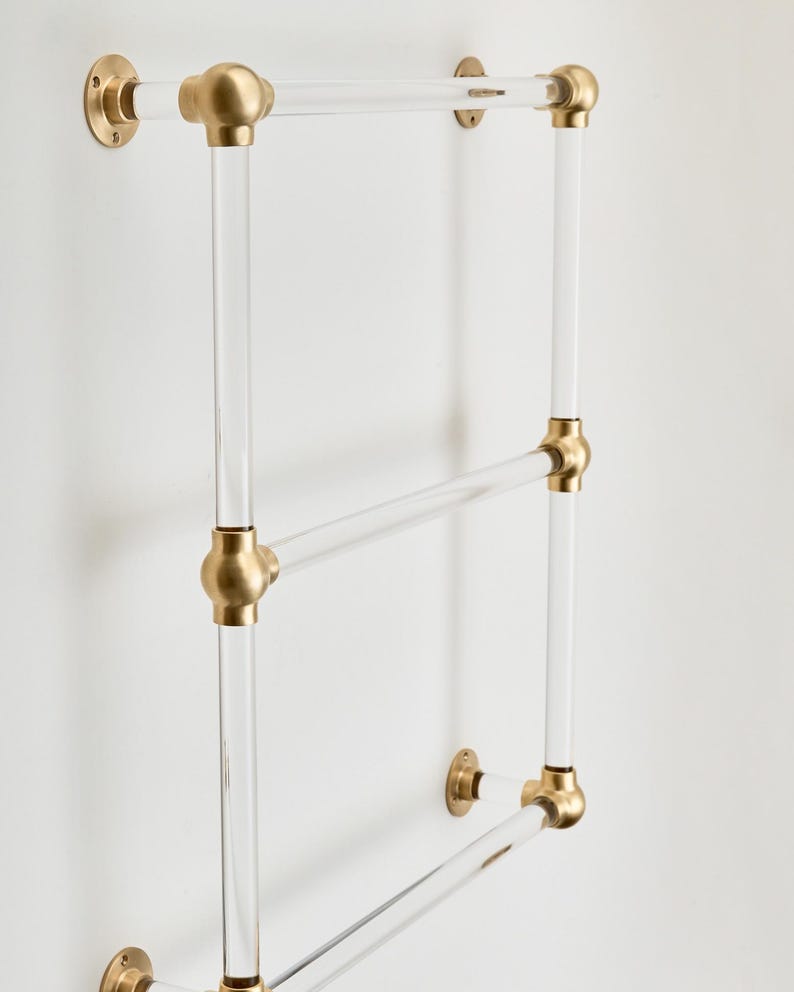 A clear glass towel rack with three horizontal bars and brass-colored hardware attached to a white wall.