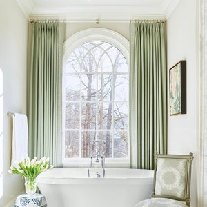 A white freestanding tub is flanked by a blue and white garden stool and a light sage green side chair. Behind the traditional tub is an arched window with a clear curtain rod installed at the top, displaying light green drapery panels.